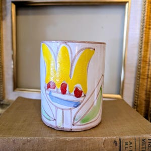May include: A cylindrical ceramic vase with a hand-painted floral design. The vase features a white base with a yellow flower, red accents, and green leaves. The vase is sitting on top of a stack of books.