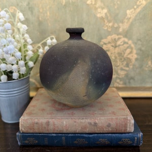 May include: A small, round, dark brown ceramic vase with a speckled glaze. The vase is sitting on top of two books, one with a floral pattern and the other a dark blue book with gold lettering.