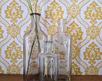 May include: Three clear glass bottles of varying sizes are arranged on a wooden surface. The bottles are in front of a yellow and white patterned wallpaper. One bottle holds purple flowers.