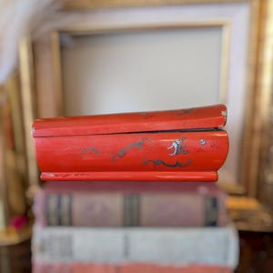 May include: A rectangular red lacquered box featuring hand-painted black and white illustrations of a dragon and other figures along the side. The box has a slightly curved lid and shows signs of wear along the edges of the vibrant red finish.