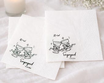 May include: White square paper napkins with a black illustration of two champagne glasses, ribbons, and the word "Engaged" with a date. The initials "R+W" are above the glasses. Ideal for a wedding or engagement celebration.