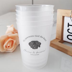 May include: A set of white plastic cups featuring a dog illustration and the text "My pawrents tied the knot! Cheers to forever! Love, Ronnie." The cups are stacked on a white surface.
