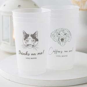 Może przedstawiać: Dwa białe plastikowe kubki z czarnymi rysunkami kota i psa. Na kubku z kotem napisano "Drinks on me!", a na kubku z psem "Coffee's on me!". Oba kubki mają prosty, elegancki design.