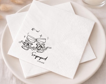 May include: White square paper napkins with a black illustration of two champagne glasses, bows, and the word "Engaged" with a date. The napkins are stacked on a white plate.