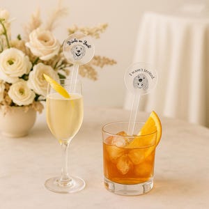 May include: Two cocktails with clear drink stirrers. One stirrer has a dog illustration and the text "Drinks on Buck!". The other stirrer has a dog illustration and the text "I wasn't invited.". One drink has a lemon peel, the other has an orange slice.