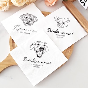 May include: White paper napkins with black line drawings of dogs. Each napkin has the phrase "Drinks on me!" in cursive, along with a dog's name and a date. The napkins are arranged on a wooden serving tray, with a floral background.