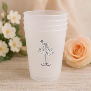 Puede incluir: Una pila de vasos de plástico esmerilado con un dibujo lineal negro de una pareja en una copa de martini. Las iniciales "J & M" están debajo del dibujo. Los vasos están sobre una superficie de madera clara, con flores en el fondo.