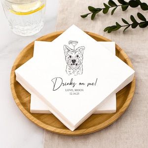 May include: White square napkins with a black line drawing of a dog with a cocktail on its head. The text "Drinks on me!" is written below the dog, along with the text "LOVE, MOON 12.16.25". The napkins are stacked on a wooden plate.