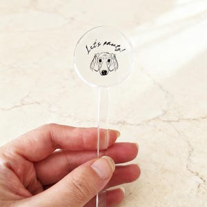 May include: A clear acrylic drink stirrer with a circular top featuring a black and white illustration of a dachshund dog and the words "Let's party!" in black script.