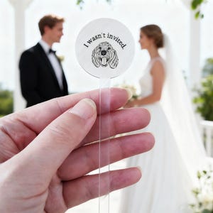May include: A round, white wedding favor with the text "I wasn't invited" and a dog illustration. The favor is held in front of a wedding scene with a bride and groom in the background. The dog illustration is a black and white line drawing.