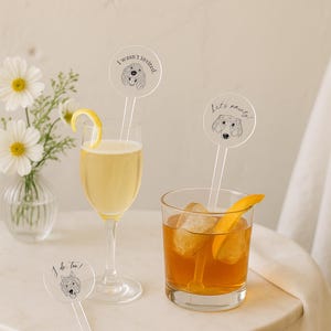 May include: Two cocktails, one in a champagne flute with a lemon twist and one in a rocks glass with ice and an orange peel. Each drink has a clear drink stirrer with a dog illustration and text: "I wasn't invited", "Let's party!", and "I do too!"