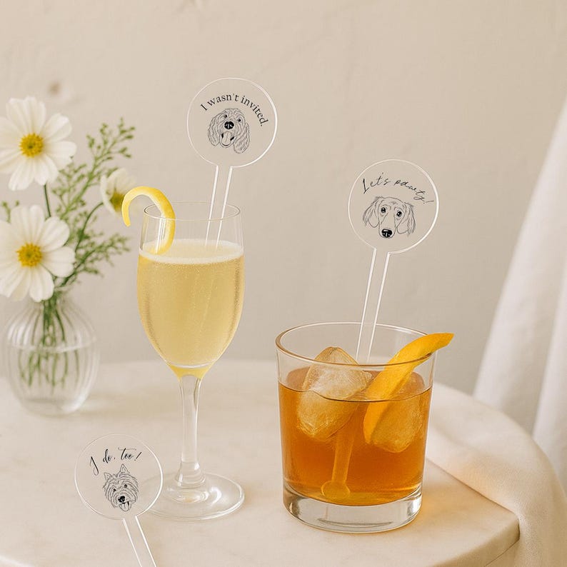 May include: Two cocktails, one in a champagne flute with a lemon twist and one in a rocks glass with ice and an orange peel. Each drink has a clear drink stirrer with a dog illustration and text: "I wasn't invited", "Let's party!", and "I do too!"