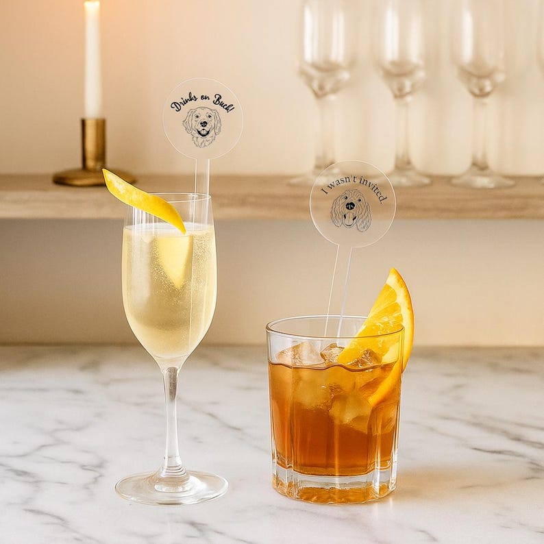 May include: Two cocktails, one in a champagne flute with a lemon twist and the other in a rocks glass with an orange slice. Clear drink stirrers feature a dog illustration and the phrases "Drinks on Buck!" and "I wasn't invited."