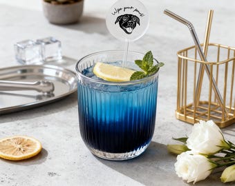 May include: A clear, ribbed glass filled with a blue gradient beverage, ice, a lemon slice, and a mint sprig. A circular acrylic drink stirrer features a black line drawing of a dog and the text "Wipe your paws!" in black script.