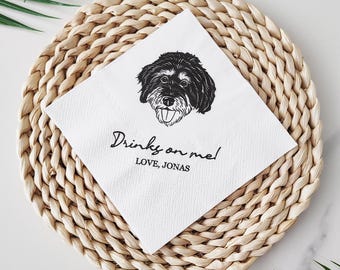 May include: A white cocktail napkin with a black and white illustration of a dog's face. The napkin has the text "Drinks on me!" and "LOVE, JONAS" in black script. The napkin is on a woven, circular placemat.