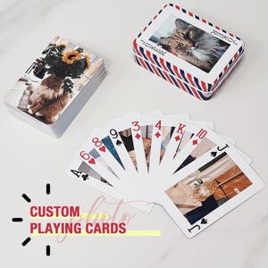 May include: Custom playing cards featuring cat photos. A deck of cards is fanned out, displaying various card suits and numbers. A stack of cards and a card box with a cat photo are also visible. The box has a red, white, and blue striped design.