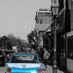 Op de afbeelding: Een vintage lichtblauwe en witte stationwagen met een chromen bumper en een kentekenplaat met de tekst "KD 359". De auto staat geparkeerd op een straat in een zwart-witte stad.