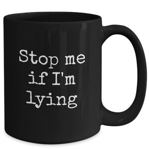 May include: A black ceramic coffee mug with a white, bold font that reads "Stop me if I'm lying". The mug has a curved handle and a glossy finish. This mug is perfect for coffee, tea, or any beverage.