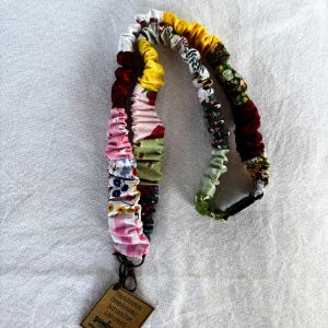 May include: A handmade scrunchie lanyard with a variety of colorful fabric patterns. The lanyard features a mix of floral, gingham, and solid-colored scrunchies in shades of pink, green, yellow, and burgundy. It includes a metal clasp and a tag that reads "Handmade Breakaway Scrunchie Lanyard."