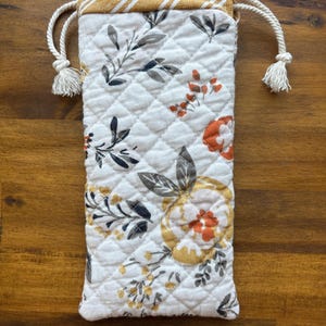 May include: A rectangular quilted drawstring pouch featuring a white base with a floral pattern in shades of orange, yellow, and gray. The top edge is a mustard yellow fabric with white diagonal stripes, finished with two white twisted rope drawstrings.