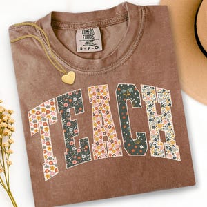 May include: A brown Comfort Colors t-shirt with the word "TEACH" in a floral design. The shirt has a crew neck and short sleeves. A gold heart pendant necklace is draped over the shirt.