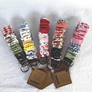 May include: Five handmade scrunchie keychain wristlets in various floral and patterned fabrics. Each wristlet has a metal ring and a brown tag that reads "Handmade Scrunchie Keychain Wristlet" and "SCHEDEN CO."
