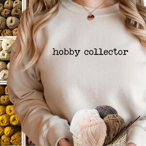 May include: A beige sweatshirt with the text "hobby collector" in black. The person is holding a small basket with yarn in various colors, including white, pink, and brown. Shelves filled with yarn are visible in the background.