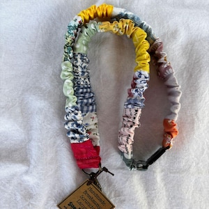 May include: A handmade breakaway scrunchie lanyard in a patchwork design. The scrunchie features various fabric patterns in colors like red, blue, yellow, and green. A small tag with text hangs from the lanyard.