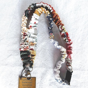 May include: A handmade breakaway scrunchie lanyard with a variety of floral and patterned fabric sections. The lanyard includes a metal clasp and a small, rectangular tag with the product name. The scrunchie fabric sections feature red, white, yellow, and blue colors.