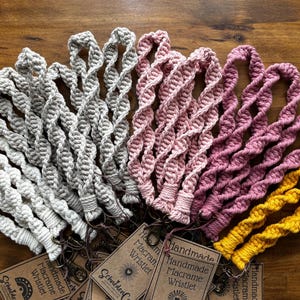 May include: A collection of macrame wristlets in white, light gray, dusty rose, and mustard yellow. Each wristlet features a twisted cord design with a metal lobster clasp and a brown paper tag labeled Handmade Macrame Wristlet.