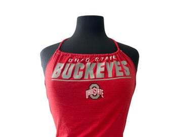 Puede incluir: Top halter rojo con el texto "OHIO STATE BUCKEYES" en letras plateadas. El logotipo de Ohio State está debajo del texto. El top se muestra en un maniquí negro.