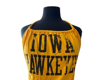 Puede incluir: Top halter mostaza con las palabras "IOWA HAWKEYES" impresas en negro. La parte superior se muestra en un maniquí negro y está hecha de un material suave, posiblemente algodón.