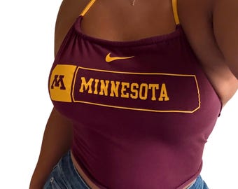 Puede incluir: Top halter burdeos con ribete dorado y la palabra "MINNESOTA" en letras doradas. El top presenta el logo de Nike y la letra "M" en un recuadro dorado. Se combina con vaqueros azules.