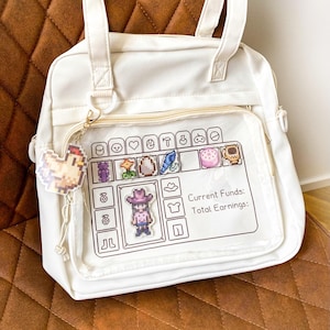 Peut inclure: Sac fourre-tout blanc avec une poche avant transparente affichant des icônes et du texte de jeu pixélisés. Un petit porte-clés poulet est suspendu à la fermeture éclair. Le sac a des poignées blanches et une fermeture éclair dorée.