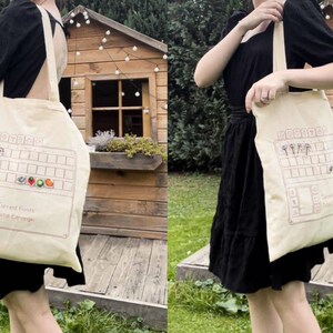 May include: A beige tote bag with a design of a video game inventory screen. The screen shows a grid of slots with icons of various items, including a mushroom, a heart, a strawberry, a tomato, and a peach. The text "Current Funds" and "Total Earnings" is visible on the screen.