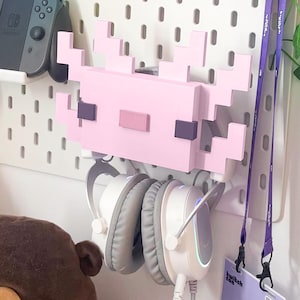 Könnte beinhalten: Ein rosa, pixeliger Axolotl-Wandhalter, der weiße und graue Kopfhörer hält. Die Halterung ist an einer weißen Lochwand befestigt, links ist ein Nintendo Switch-Controller zu sehen. Ein lila Schlüsselband mit einem TwitchCon-Abzeichen hängt rechts.