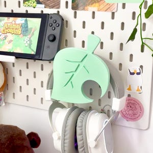 Könnte beinhalten: Eine weiße Lochwand mit einem mintgrünen Kopfhörerhalter in Blattform, der weiße und graue Kopfhörer hält. Eine Nintendo Switch mit dem Spiel Animal Crossing ist sichtbar. Die Lochwand hat auch dekorative Karten und Aufkleber.