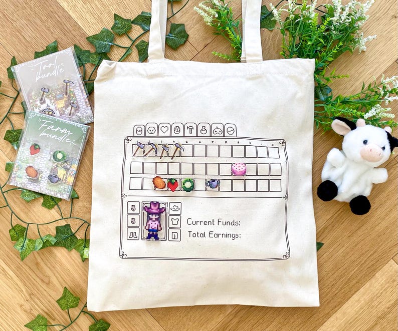 May include: A white canvas tote bag with a black and white design of a video game-style farm. The design includes a grid with icons for tools, crops, and a character. The text "Current Funds: Total Earnings:" is also included in the design. The bag is surrounded by green plants and a white stuffed cow.