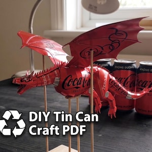 Op de afbeelding: Een draaksculptuur gemaakt van rode en zwarte Coca-Cola Zero Sugar aluminium blikjes, met gedetailleerde vleugels, een stekelige rug en een lange staart. De figuur wordt ondersteund door dunne houten stokjes. Tekst luidt: DIY Tin Can Craft PDF.
