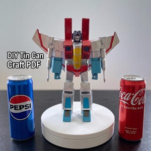 Puede incluir: Una figura de robot construida con latas de metal, con paneles rojos, plateados y azules y una pieza amarilla en el pecho. La figura se encuentra sobre una base circular blanca entre una lata de Pepsi azul y una lata de Coca-Cola roja.