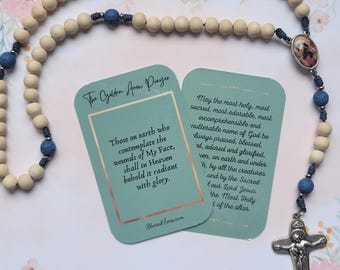 May include: A wooden rosary with blue beads and a silver crucifix. The rosary is accompanied by two cards with text about the Golden Arrow Prayer. The text on the cards reads: "Those on earth who contemplate the wounds of My Face, shall in Heaven behold it radiant with glory." and "May the most holy, most sacred, most adorable, most incomprehensible and nutterable name of God be always praised, blessed, adored and glorified, even, on earth and under heaven, by all the creatures and by the Sacred Heart of our Lord Jesus the Most Holy of the altar."