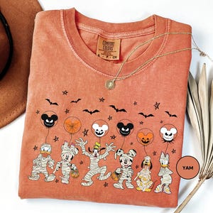 a t-shirt with a Halloween-themed design, including characters such as Mickey Mouse, Minnie Mouse, Donald Duck, and other spooky and festive figures. The t-shirt is displayed on a white background, with a brown hat and a fan visible in the background.