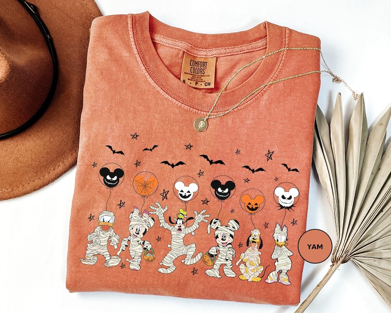 a t-shirt with a Halloween-themed design, including characters such as Mickey Mouse, Minnie Mouse, Donald Duck, and other spooky and festive figures. The t-shirt is displayed on a white background, with a brown hat and a fan visible in the background.