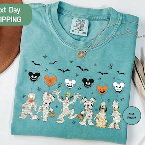 A turquoise t-shirt with a Halloween-themed design featuring characters such as Mickey Mouse, Donald Duck, Goofy, and Pluto, along with bats, pumpkins, and a sea foam design.