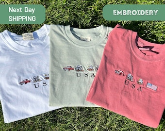 Può includere: Tre magliette ricamate con un camioncino rosso, aquile e la parola "USA". Le magliette sono bianche, verde chiaro e rosa.