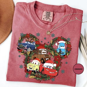 A pink t-shirt with a Christmas-themed design featuring cartoon cars and characters, including a Santa hat, a blue car, and a red car.