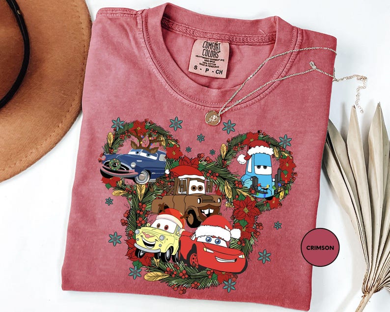 A pink t-shirt with a Christmas-themed design featuring cartoon cars and characters, including a Santa hat, a blue car, and a red car.