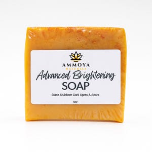 Może przedstawiać: Kwadratowy blok mydła Advanced Brightening, zapakowany w przezroczysty plastik. Mydło ma ciepły pomarańczowy kolor. Biała etykieta zawiera nazwę marki, logo kwiatu lotosu oraz napis "Advanced Brightening SOAP". Na etykiecie podano również, że mydło waży 113 gramów.