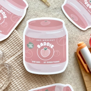 May include: Pink and white sticker with a peach design and the text "Pre-Workout Baddie" and "She bad... at everything".