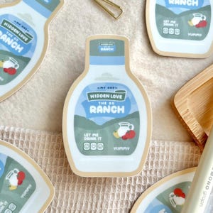 May include: A collection of stickers shaped like ranch dressing bottles. Each sticker features the text "Hidden Love The OG Ranch" in blue and white, along with a graphic of a pitcher and fruit. The phrase "Let Me Drink It" is also included.